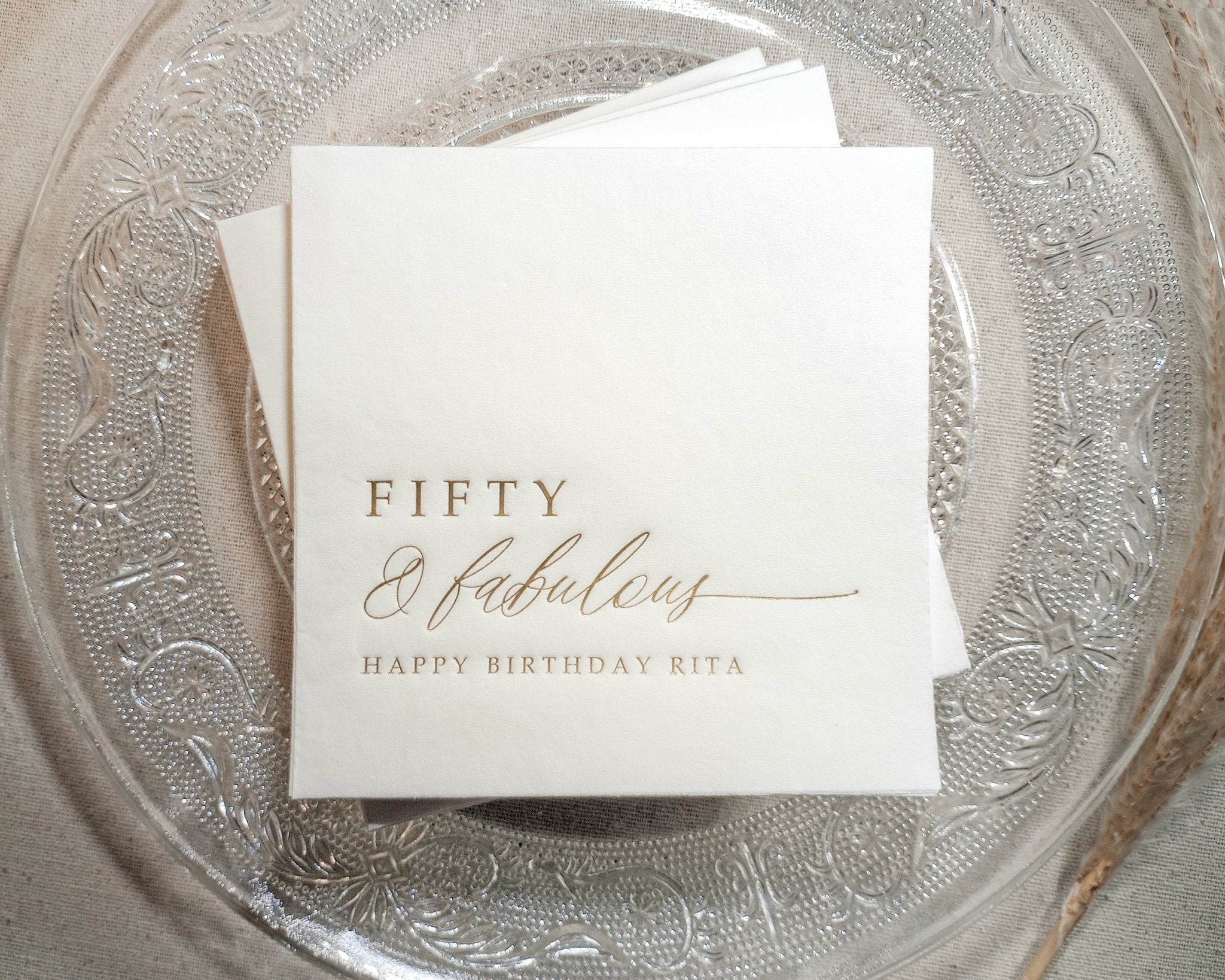 Two personalized birthday napkins with gold foil on charger plate, styled for elegant party setup