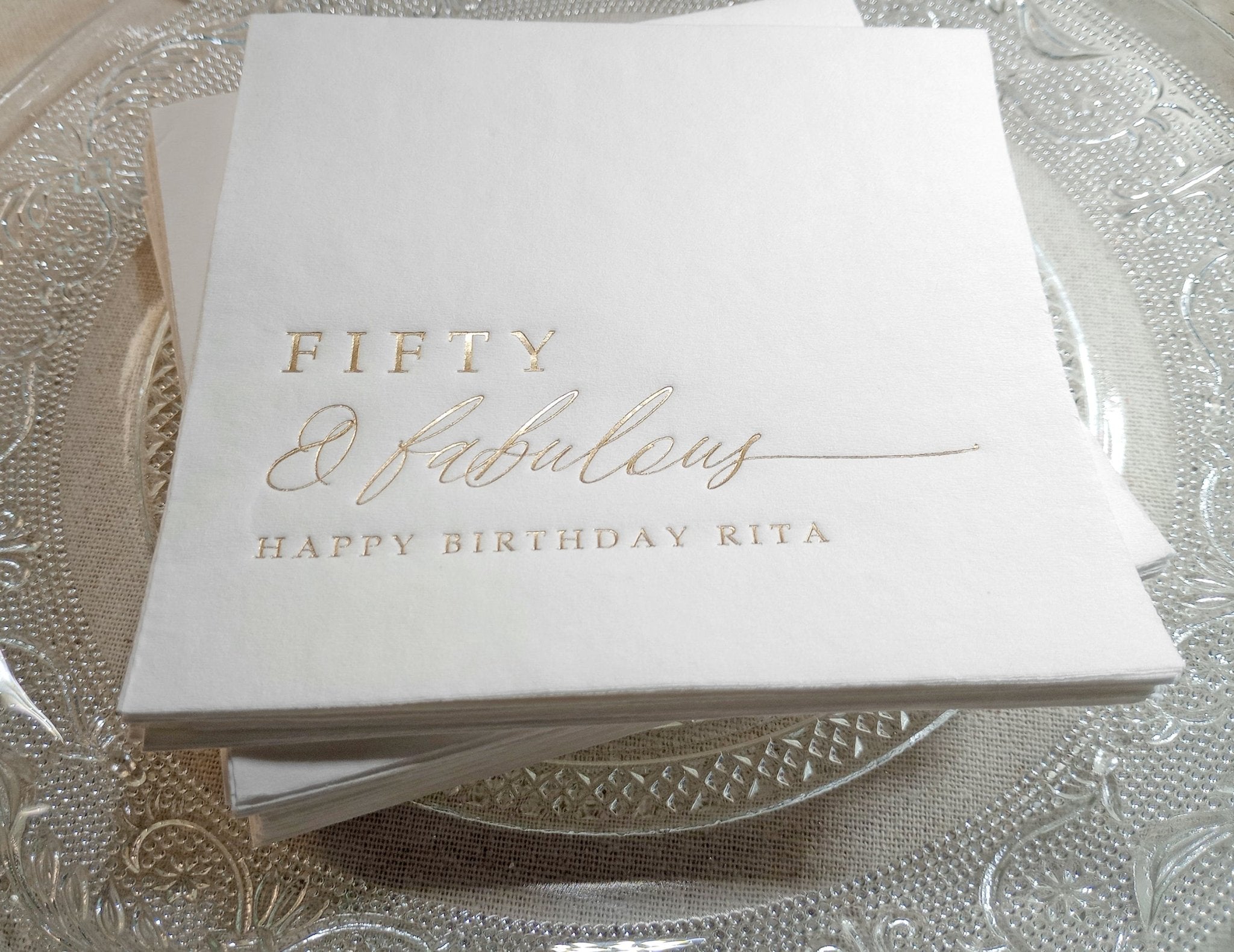 Gold foil “Fifty & Fabulous” birthday napkin with personalized name on clear glass plate