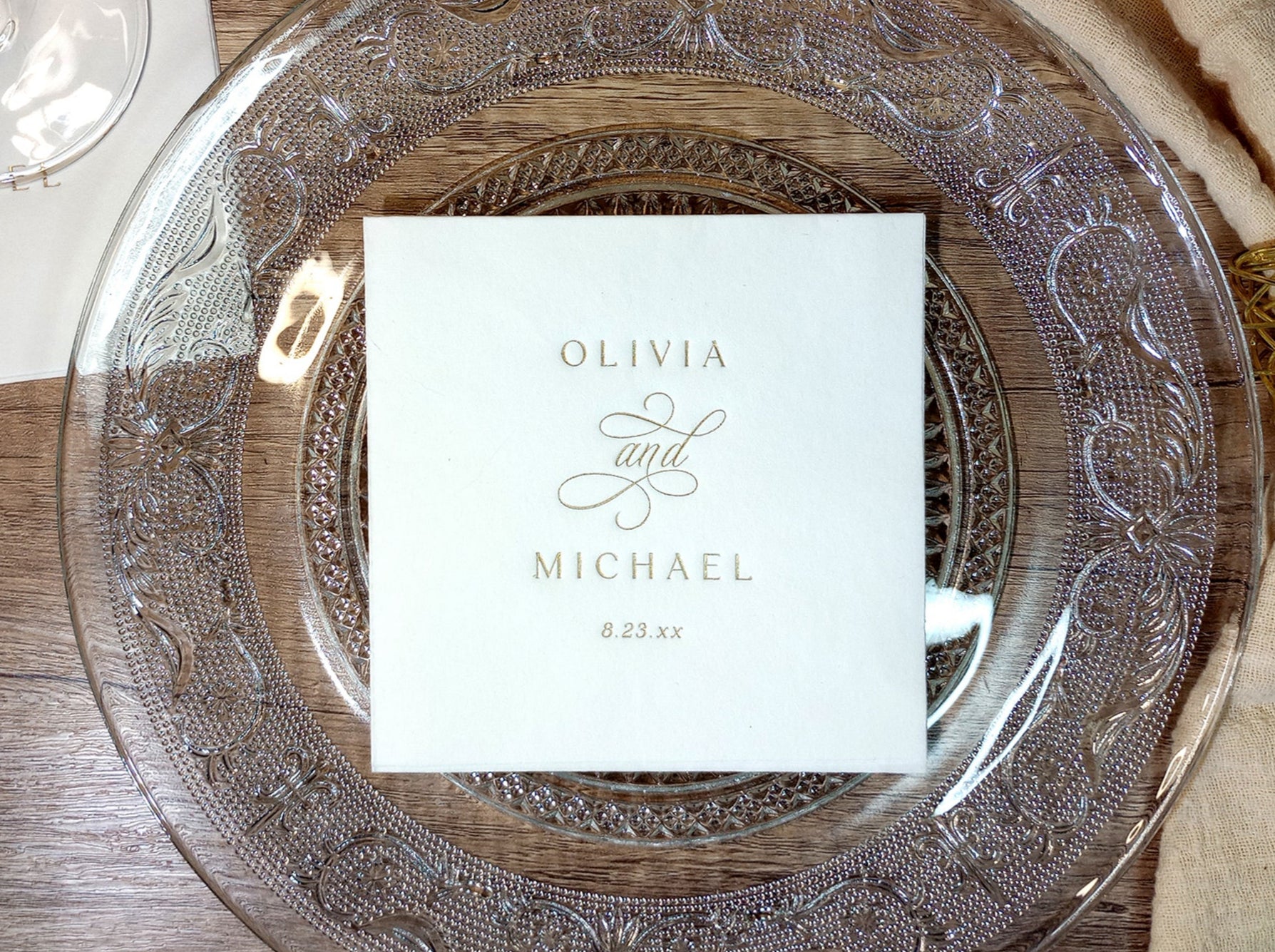 Vintage-style custom wedding napkin with elegant monogram design, shown on a glass charger plate with gold foil stamp.