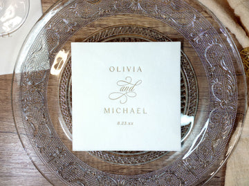 Vintage-style custom wedding napkin with elegant monogram design, shown on a glass charger plate with gold foil stamp.