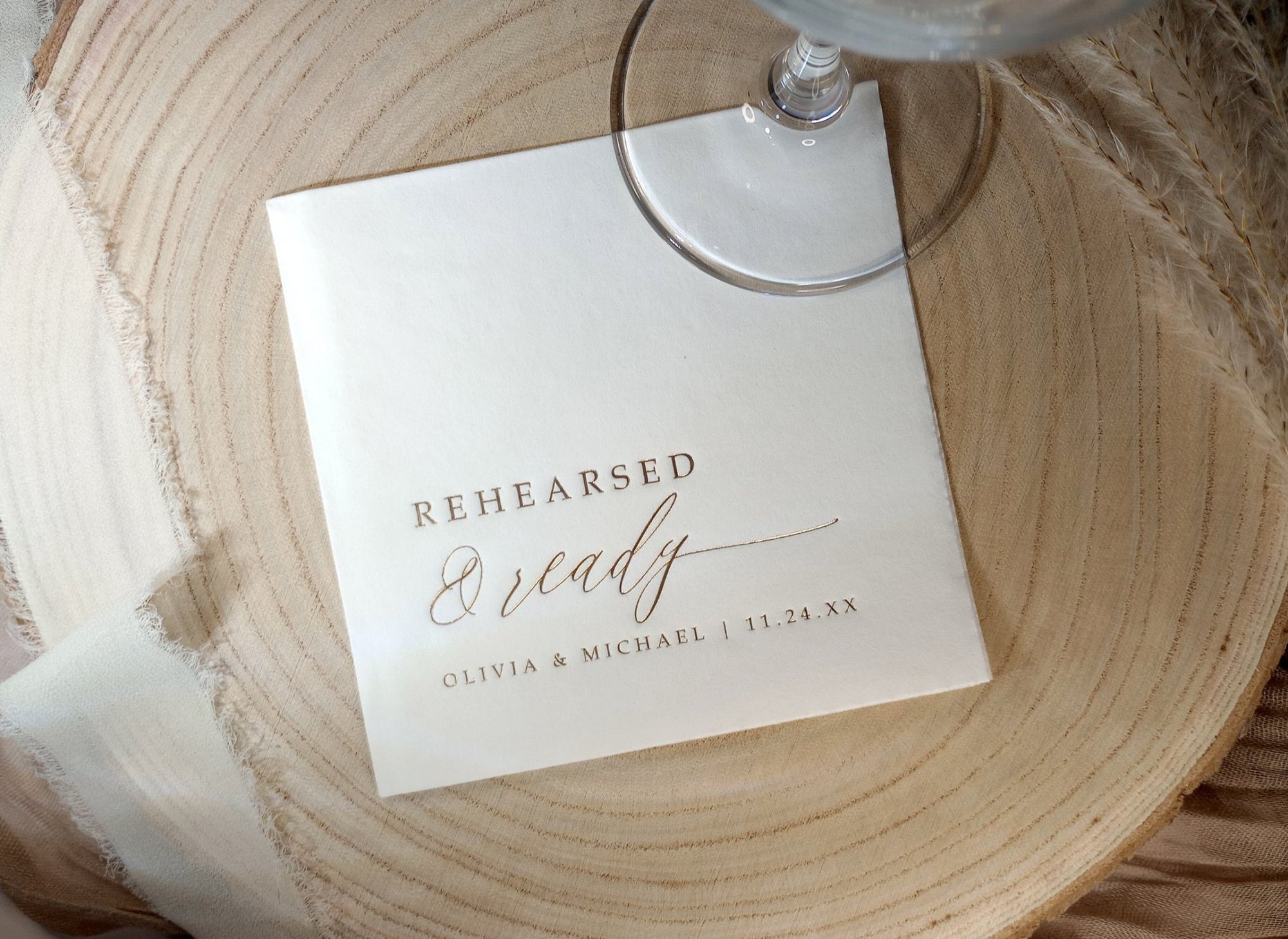 White cocktail napkin with “Rehearsed & Ready” in gold foil beside wine glass on wood charger