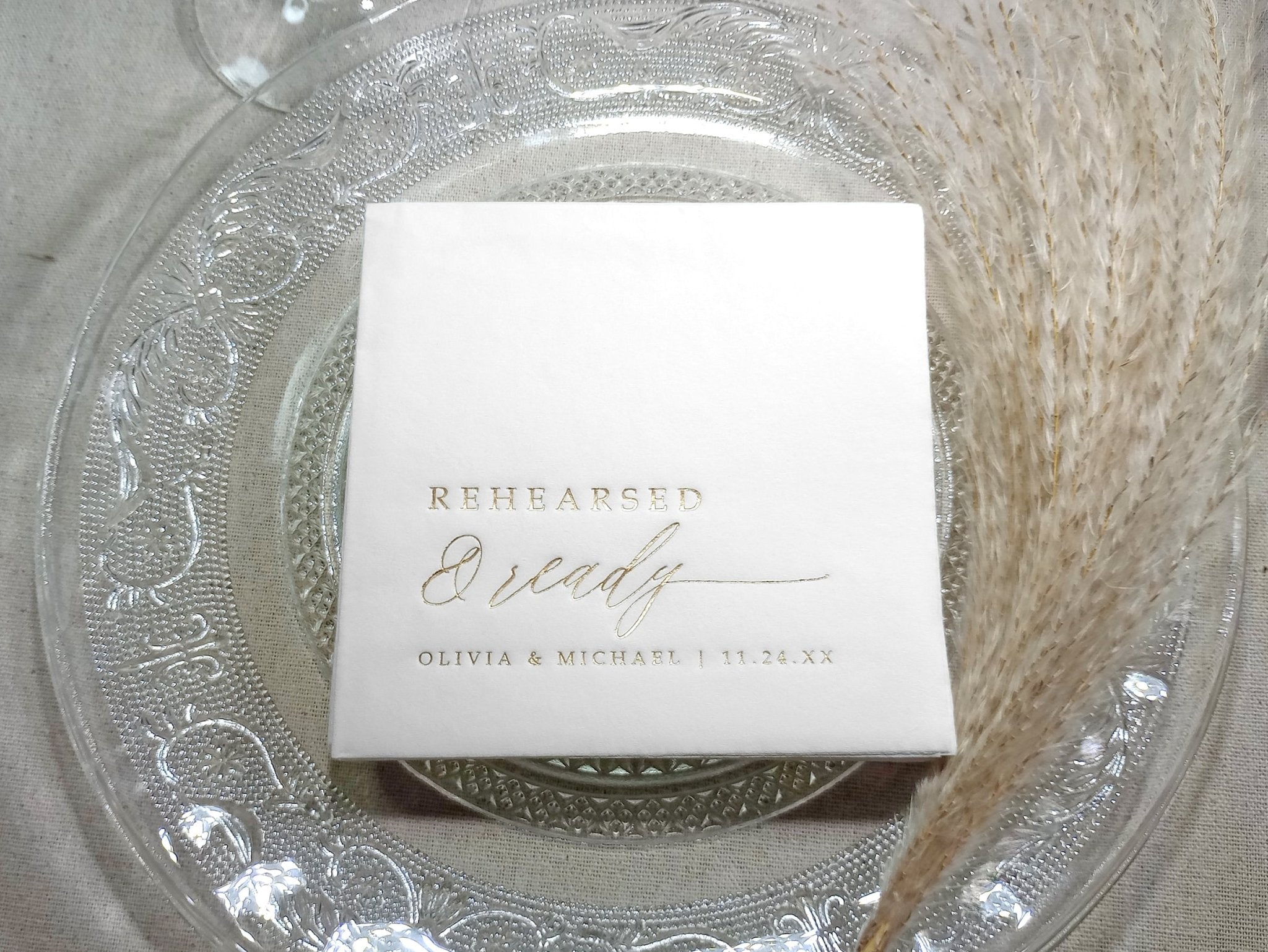 White rehearsal dinner napkins with 'Rehearsed & Ready' text on a decorative glass plate.