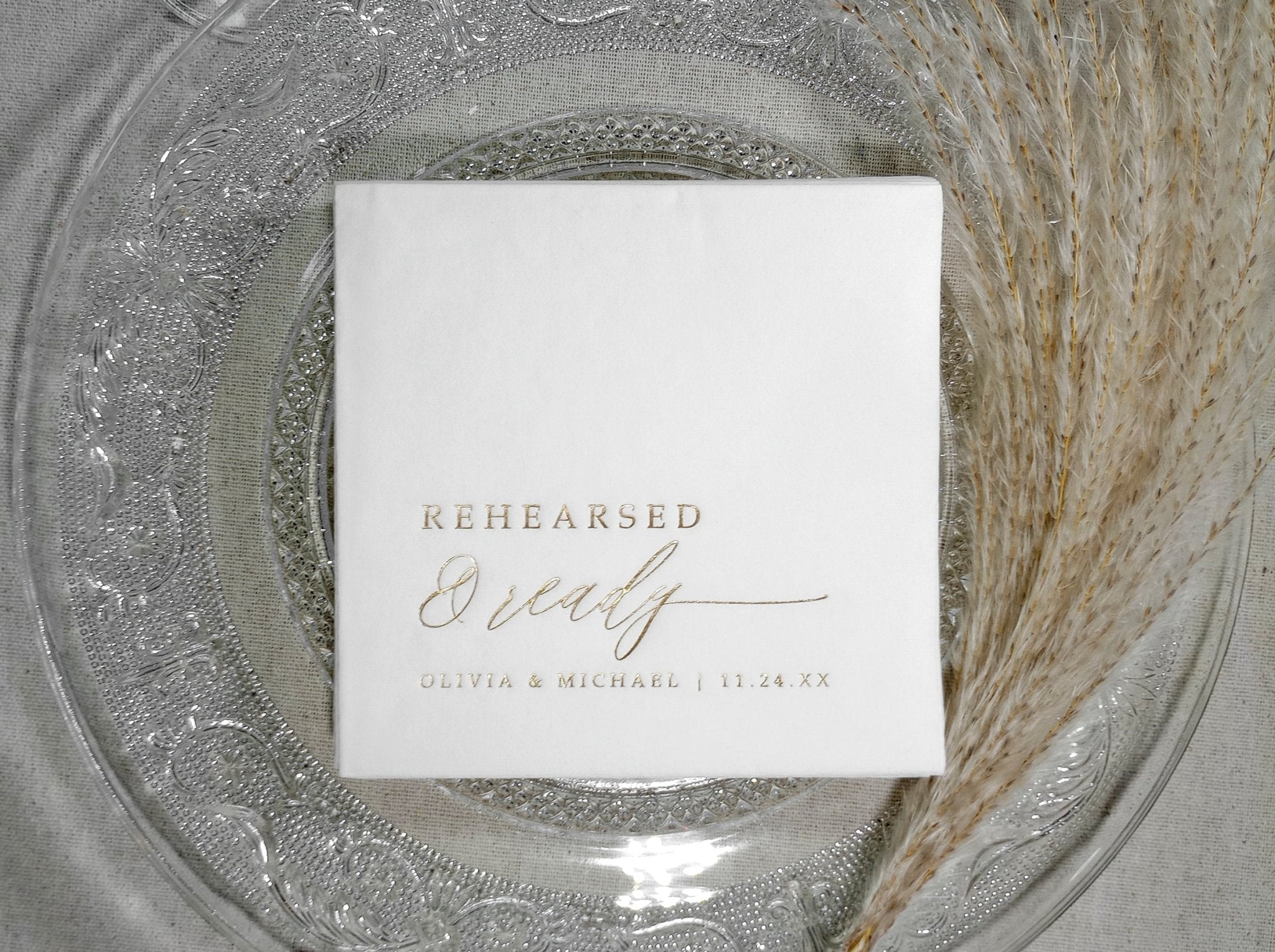 White rehearsal dinner napkin with 'Rehearsed & Ready' text on a decorative glass plate.
