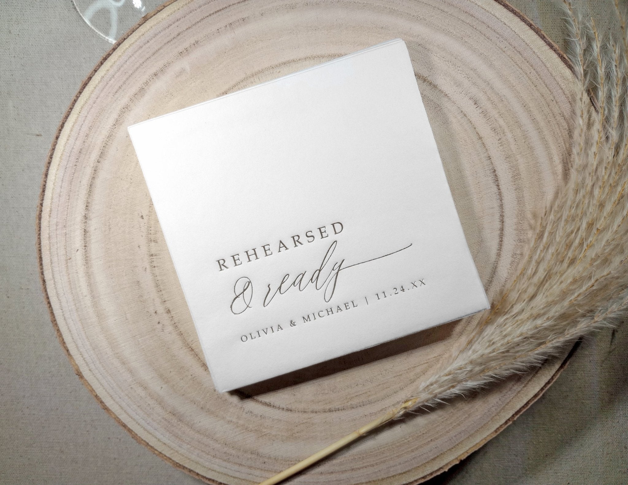 White rehearsal dinner napkins with silver foil 'Rehearsed & Ready' text on a wooden surface