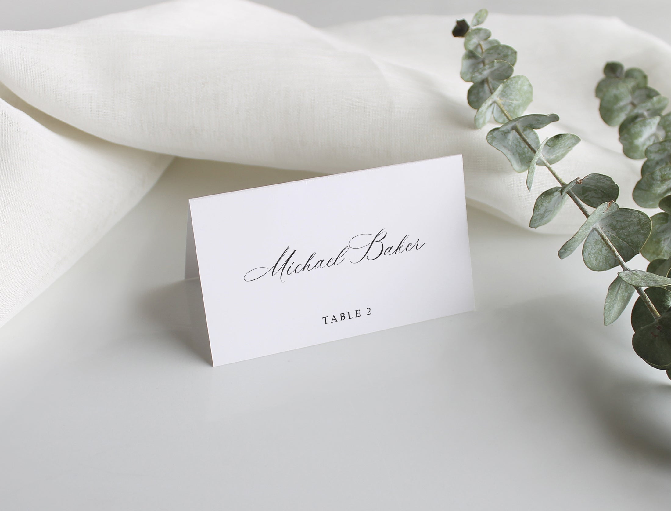 Classic wedding place card with guest name and table number, displayed with soft linens and eucalyptus accent.