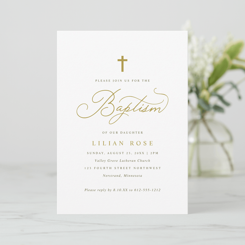 Baptism invitation card with gold text on a white background, blurred flowers in the background.