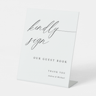 Elegant calligraphy guest book sign reading “Kindly Sign,” styled with floral wedding décor.