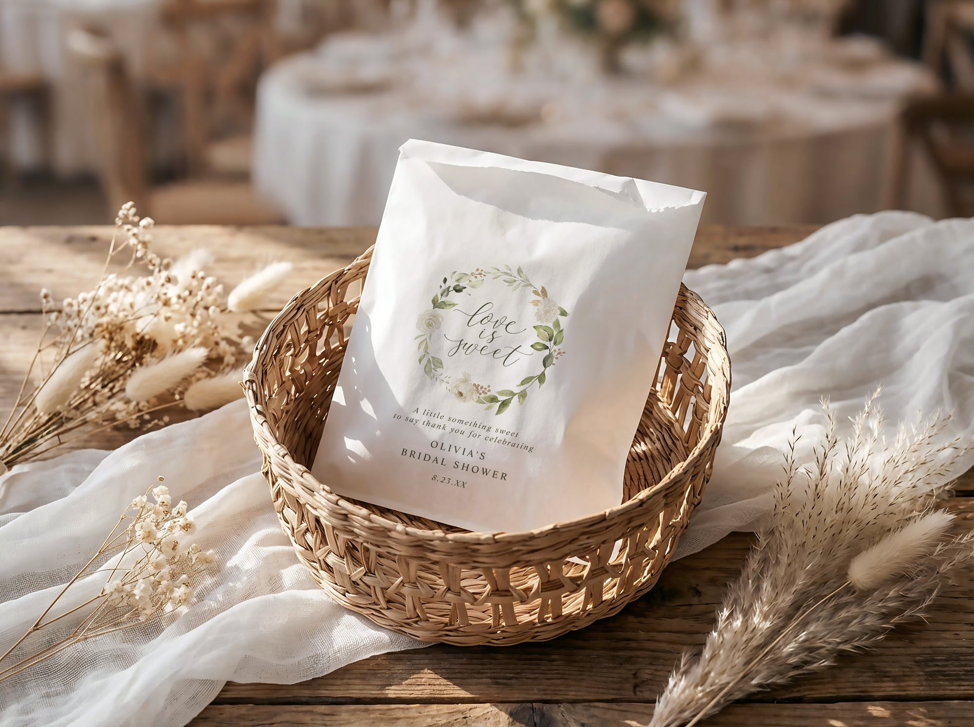 White bridal shower favor bag with greenery design in a woven basket on a wooden table with white fabric and dried plants.