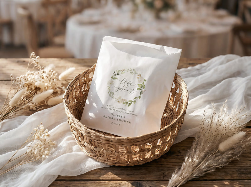 White bridal shower favor bag with greenery design in a woven basket on a wooden table with white fabric and dried plants.