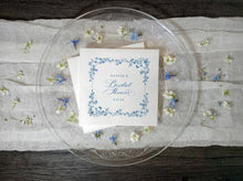 White bridal shower napkins with a blue vintage floral border and elegant script text, displayed on a clear glass plate over a white fabric runner with scattered small flowers on a dark wood surface.