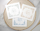 Three personalized bridal shower napkins with vintage floral borders and elegant script text, arranged on a round natural wood slice with soft white fabric and dried grass props.