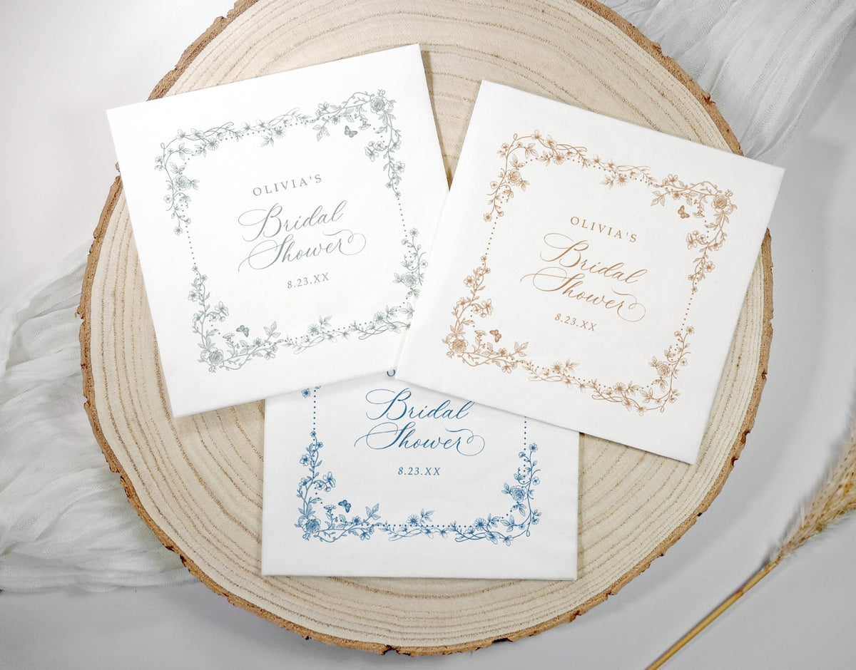Three personalized bridal shower napkins with vintage floral borders and elegant script text, arranged on a round natural wood slice with soft white fabric and dried grass props.