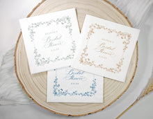 Three personalized bridal shower napkins with vintage floral borders and elegant script text, arranged on a round natural wood slice with soft white fabric and dried grass props.