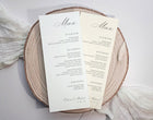 Two elegant wedding menu cards custom printed with black script and serif text on cream paper, displayed on a round natural wood slice coaster with soft white fabric in the background.