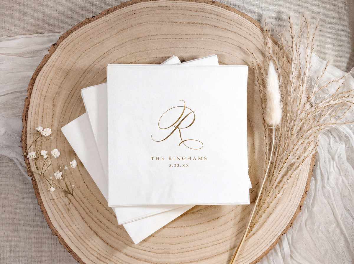 A stack of white paper napkins with a gold monogram logo and the text 'THE RINGHAMS 8.25 AM' on the top napkin, arranged on a round wood slice with dried grass stems and small white flowers.