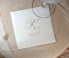 A square white paper wedding napkin with a large cursive monogram and the text 'THE KAISERS' and 'December 12, 2022,' placed on a round wood slice with part of a glass stem visible at the top.