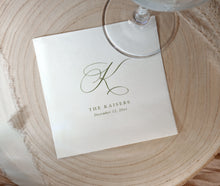 A square white paper wedding napkin with a large cursive monogram and the text 'THE KAISERS' and 'December 12, 2022,' placed on a round wood slice with part of a glass stem visible at the top.