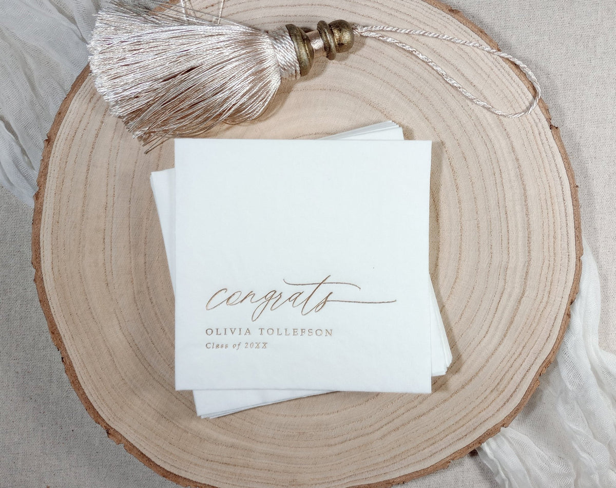 A stack of white paper personalized graduation napkins printed with elegant cursive text reading 'congrats OLIVIA TOLLEFSON Class of 20xx,' styled on a round natural wood slice with a metallic tassel charm resting above.