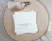 A stack of white paper personalized graduation napkins printed with elegant cursive text reading 'congrats OLIVIA TOLLEFSON Class of 20xx,' styled on a round natural wood slice with a metallic tassel charm resting above.
