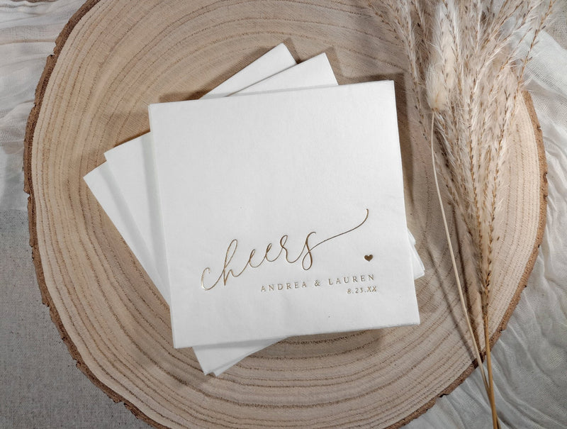 Stack of white napkins with 'cheers' and personalized text on a wooden surface