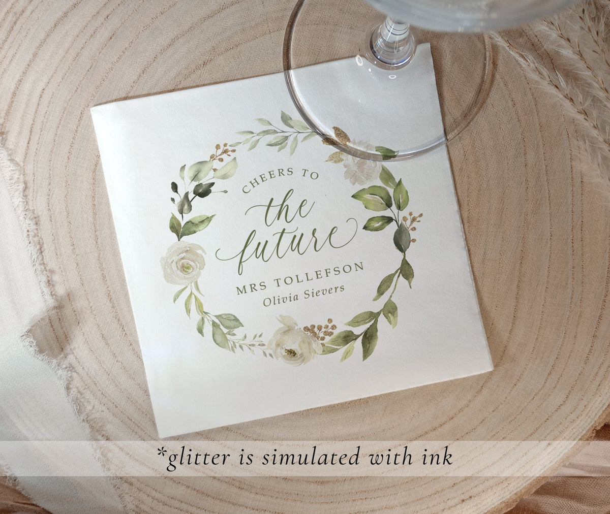 A printed bridal shower napkin with a floral wreath design and the text 'Cheers to the future Mrs Tollefson Olivia Siemers,' placed on a round wood slice with part of a glass stem visible. A translucent banner at the bottom says glitter is simulated with ink.