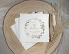 A stack of white bridal shower napkins with a watercolor floral wreath and the text 'Cheers to the future Mrs Tollefson' is styled on a round wood slice with dried grasses.
