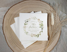 A stack of white bridal shower napkins with a watercolor floral wreath and the text 'Cheers to the future Mrs Tollefson' is styled on a round wood slice with dried grasses.