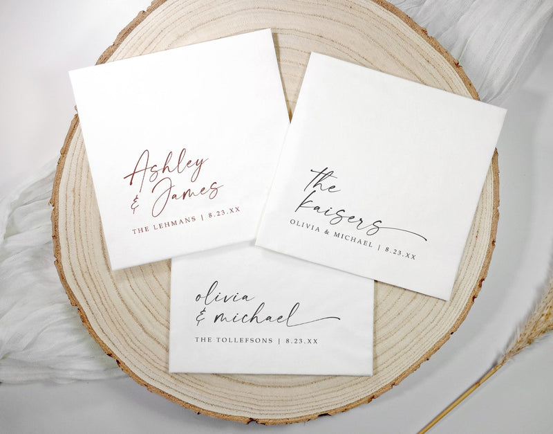 Three white wedding napkins with elegant handwriting script names and serif typography, arranged on a round natural wood slice coaster over a white fabric background.