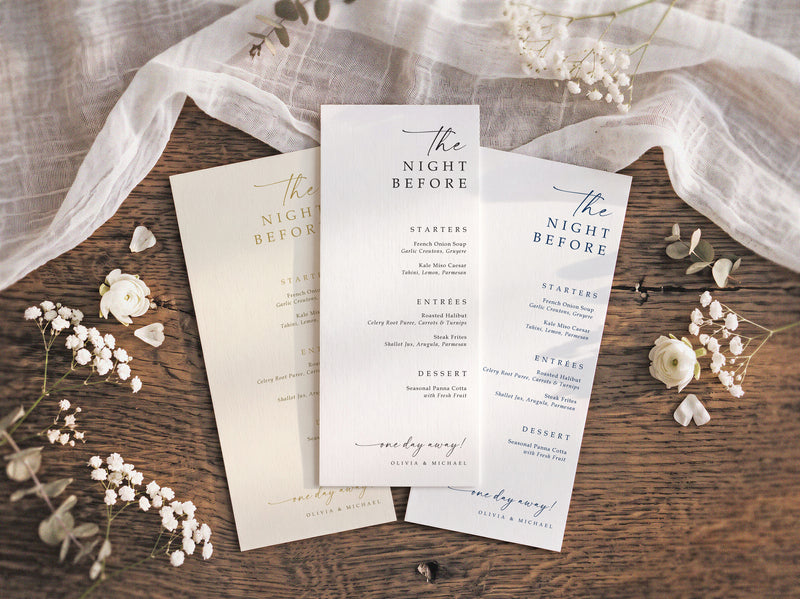 Three wedding rehearsal dinner menu cards for 'The Night Before' arranged on a rustic wooden table with white fabric and small white flowers.