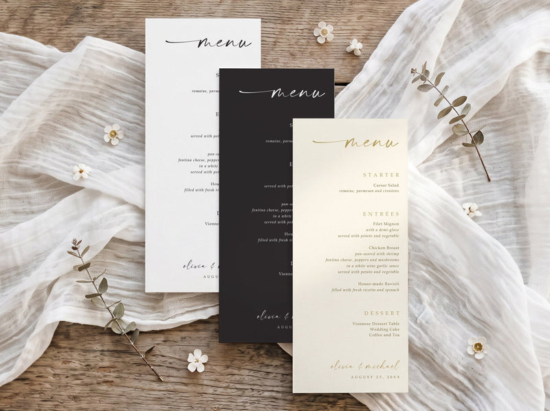 Three elegant printed wedding menu cards arranged in a flat lay on a rustic wooden surface with draped white fabric and small floral accents. The set includes one white card with black script, one black card with white script, and one cream card with gold script.