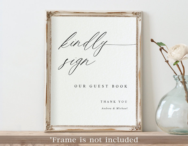 Framed sign with 'kindly sign our guest book' text on a white wall with a vase of flowers.