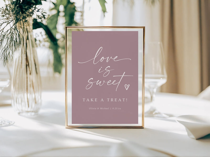 Framed sign on a table with 'love is sweet' text, surrounded by greenery and white tablecloth.