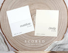Two white name cards on a wooden surface with text 'matthew evans' and 'emily baker'.