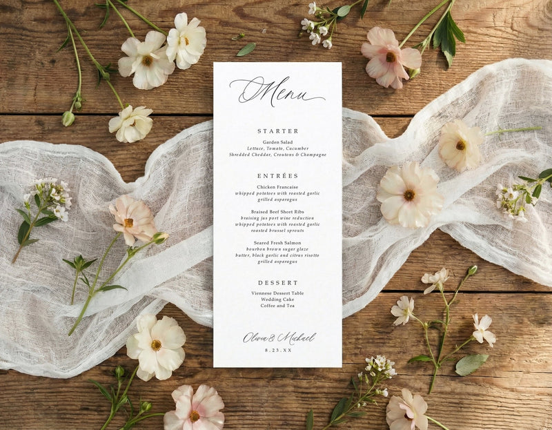 A vertical printed wedding menu card with elegant script and serif text, styled in a flat lay on a rustic wooden surface with white lace fabric and scattered cream and blush flowers.