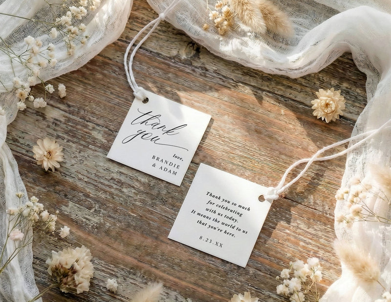 Two 'thank you' favor tags on a wooden surface with dried flowers.