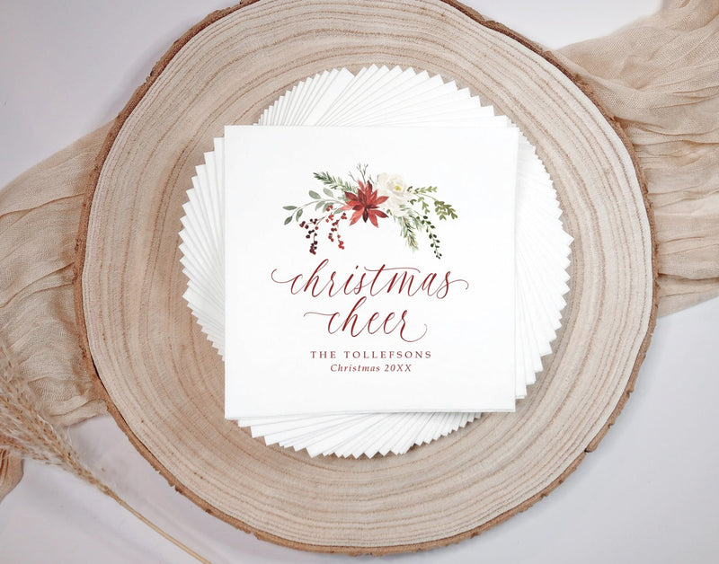 A stack of white personalized paper napkins with a watercolor Christmas floral design and the words 'christmas cheer' in red script, displayed on a round wood slice with beige fabric and dried wheat around it.