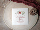 A square white Christmas cocktail napkin with a watercolor floral wreath and the words 'christmas cheer' in red script, placed on a round wood slice with a pinecone and dark red fabric around it.
