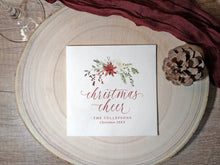 A square white Christmas cocktail napkin with a watercolor floral wreath and the words 'christmas cheer' in red script, placed on a round wood slice with a pinecone and dark red fabric around it.