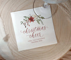 A square white printed cocktail napkin with a floral Christmas illustration and the words 'christmas cheer' in cursive, placed on a round wood slice with part of a glass visible at the top.