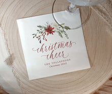 A square white printed cocktail napkin with a floral Christmas illustration and the words 'christmas cheer' in cursive, placed on a round wood slice with part of a glass visible at the top.