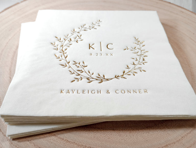 A stack of white paper napkins with a gold foil monogram wreath design and the names 'KAYLEIGH & CONNER' foil stamped on them, displayed on a round wooden surface.