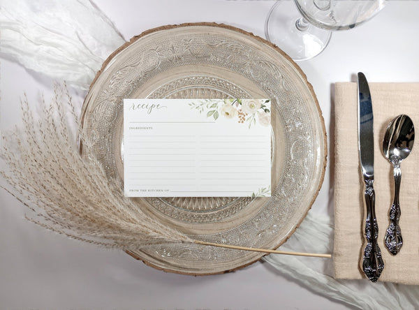Elegant table setting with a decorative plate, bridal shower recipe card with delicate watercolor florals, and cutlery on a white surface.