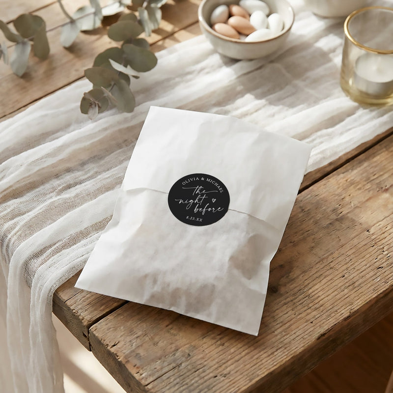 A black rehearsal dinner favor bag sticker that says "the night before" in an elegant handwriting script and is personalized with the couple's names and date.