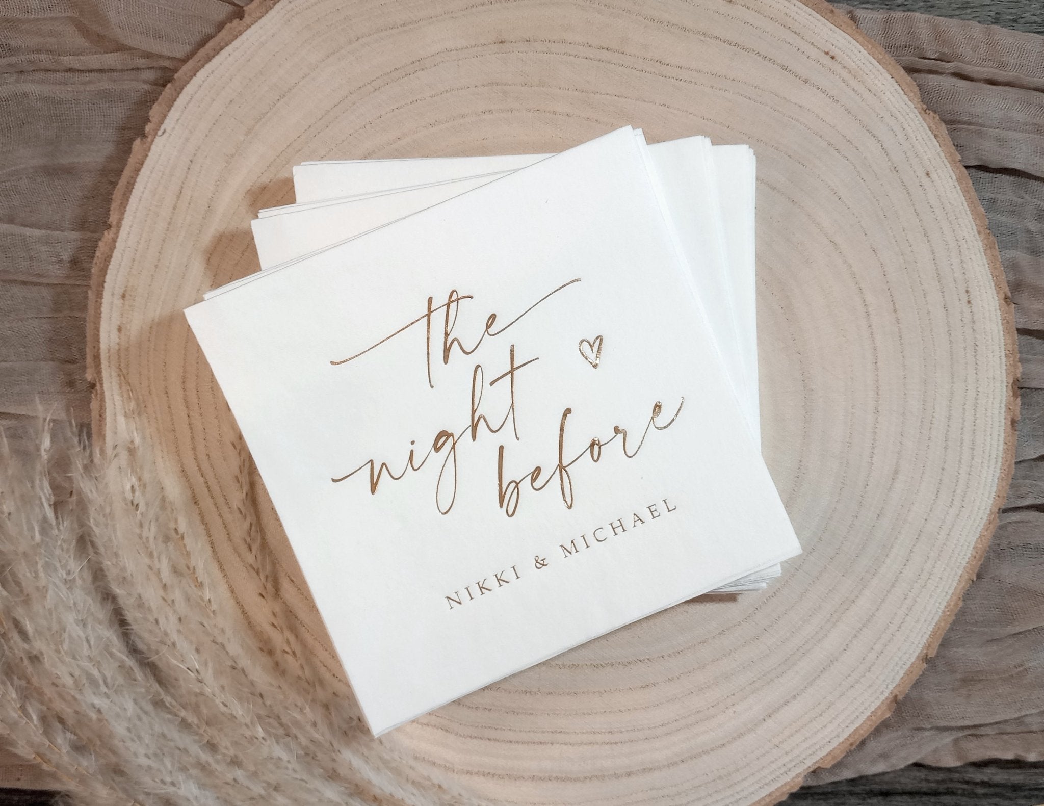 Custom rehearsal dinner napkin with ‘The Night Before’ in gold foil and personalized names, displayed on wood.