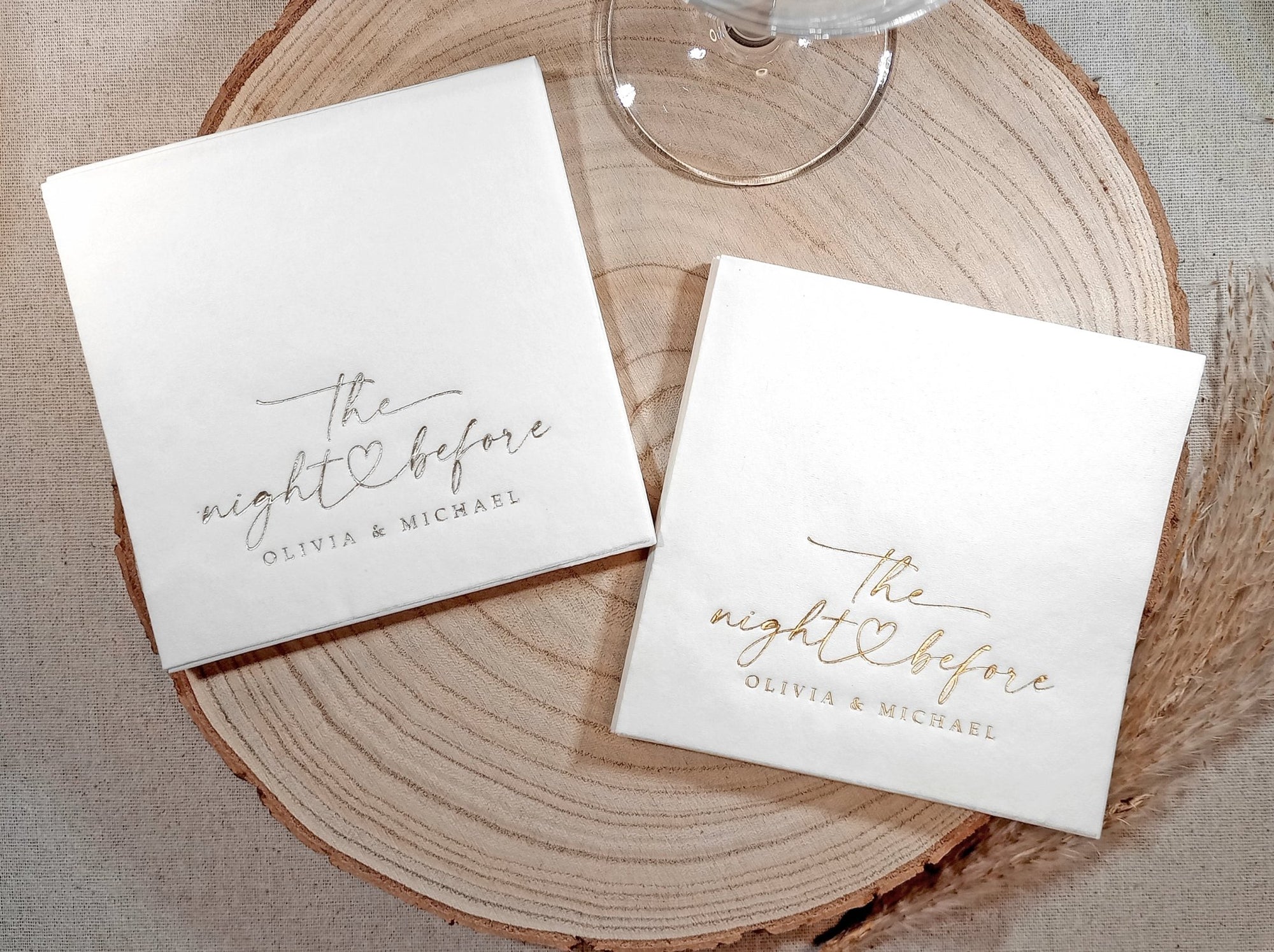 wo stacks of white napkins—one with gold foil and one with silver foil—both featuring “the night before” script with a heart and names.