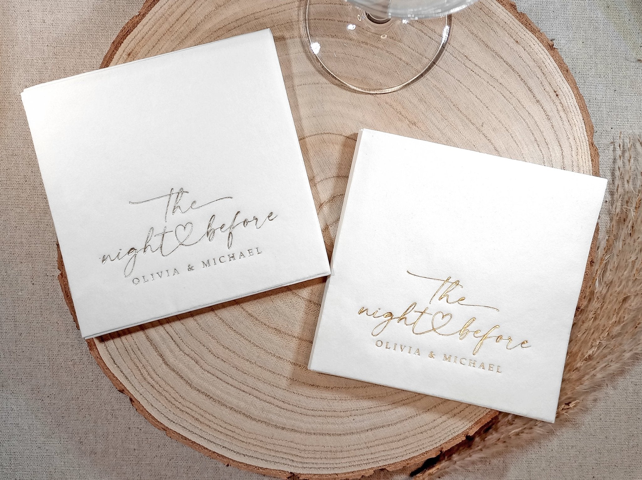 wo stacks of white napkins—one with gold foil and one with silver foil—both featuring “the night before” script with a heart and names.