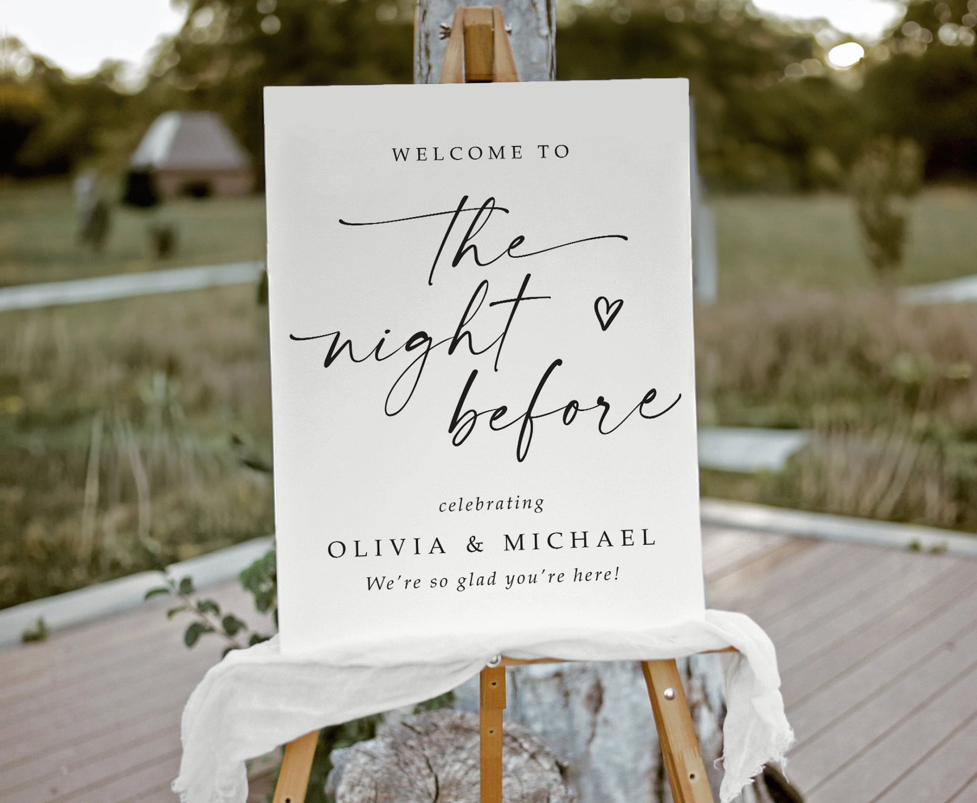 White foam board sign that says "the night before" with hand-drawn heart and custom names on a scenic background.