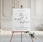 Rehearsal dinner welcome sign on an easel with floral arrangement in a room with white walls and wooden floor.