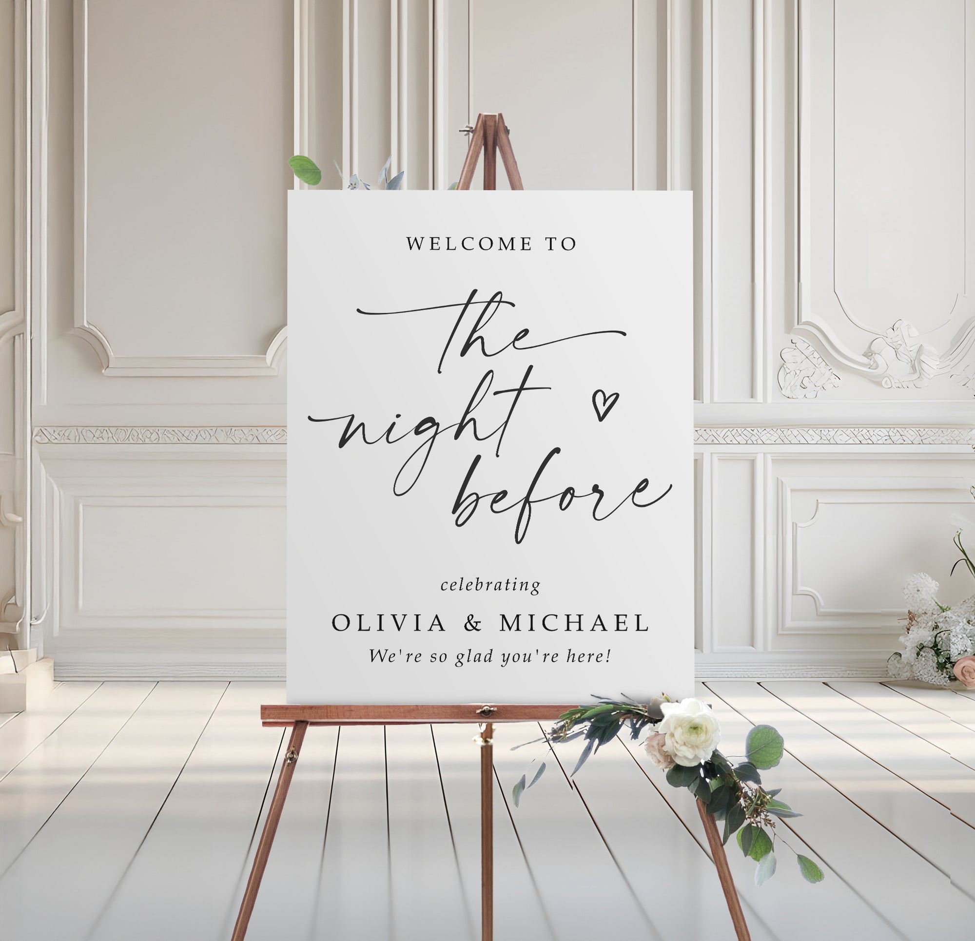 Rehearsal dinner welcome sign on an easel with floral arrangement in a room with white walls and wooden floor.
