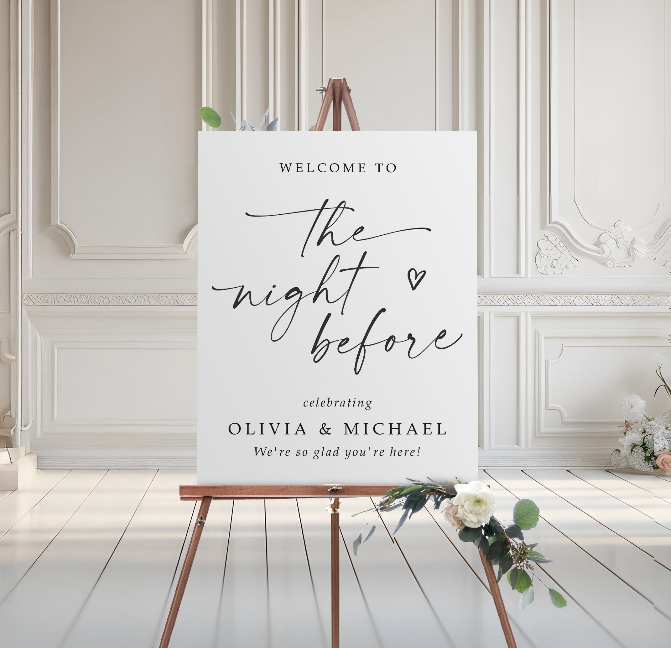 Rehearsal dinner welcome sign on an easel with floral arrangement in a room with white walls and wooden floor.